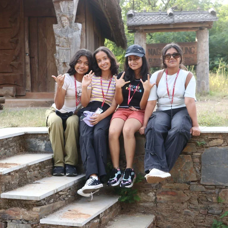 Four students sitting on stone steps making hand gestures, wearing lanyards, with traditional Naga wooden structures behind them