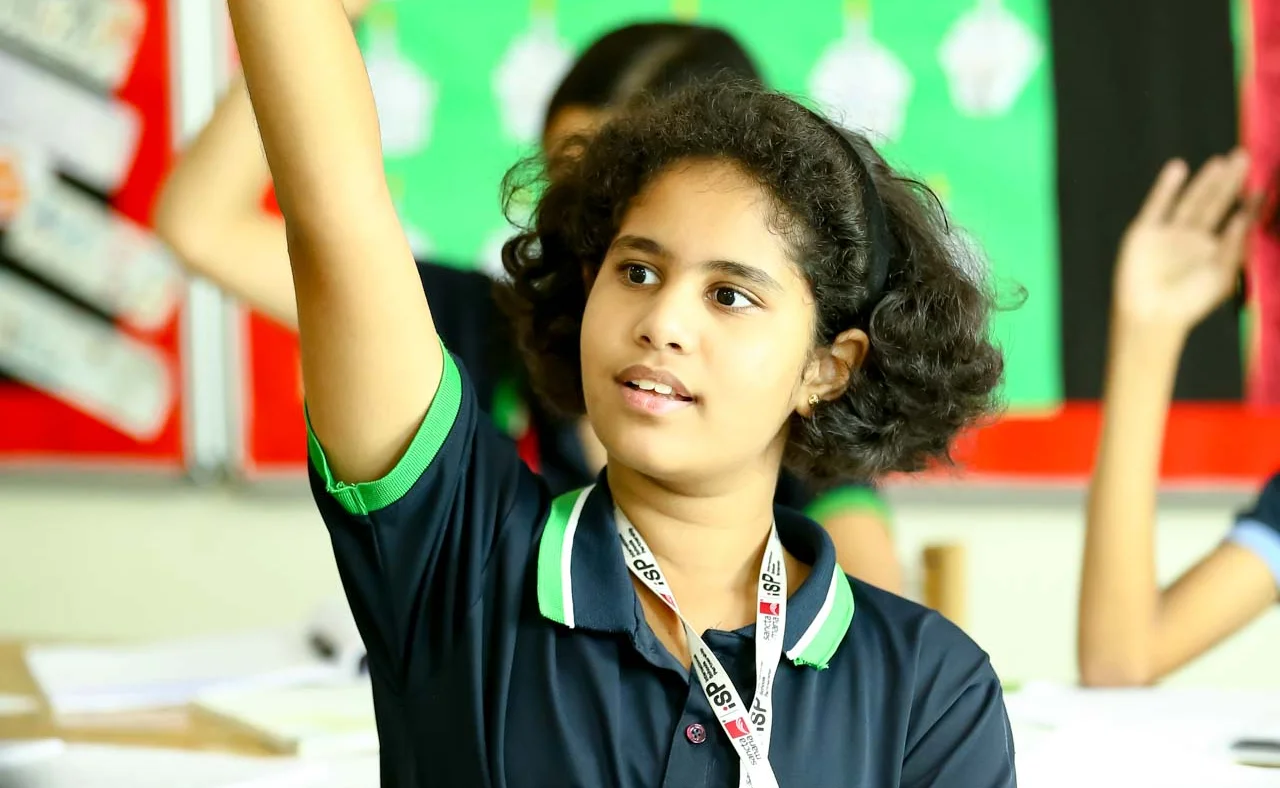 Student in Sancta Maria uniform raising hand with pen during classroom lesson, other students visible in background
