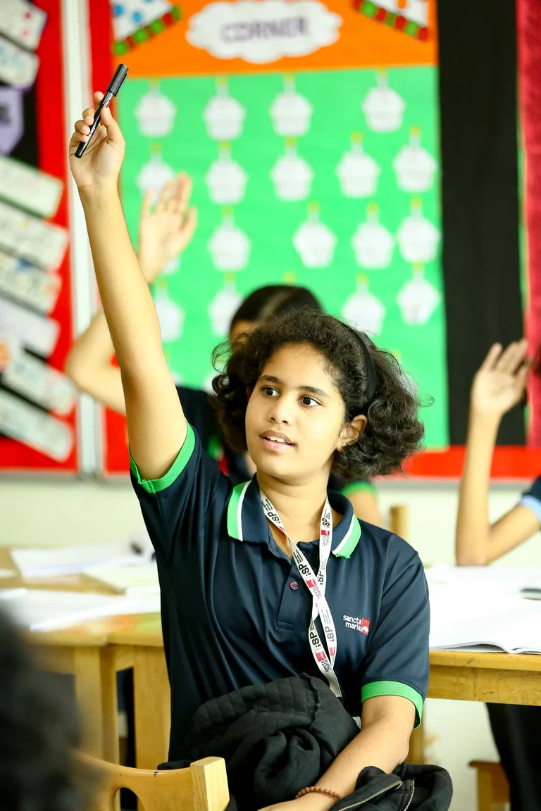 Student in Sancta Maria uniform raising hand with pen during classroom lesson, other students visible in background