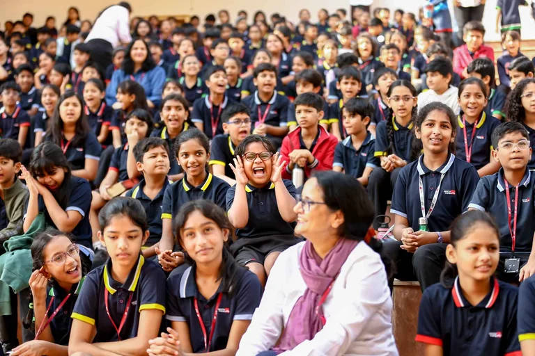 Students in navy and yellow uniforms laughing and cheering during a school assembly, with one girl excitedly cupping her hands