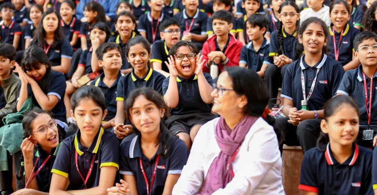 Students in navy and yellow uniforms laughing and cheering during a school assembly, with one girl excitedly cupping her hands