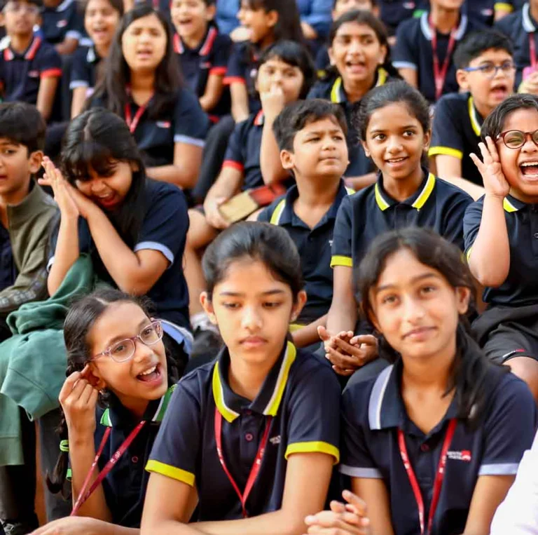 Students in navy and yellow uniforms laughing and cheering during a school assembly, with one girl excitedly cupping her hands