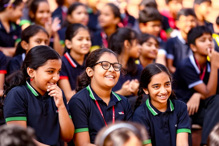 Smiling students in navy blue and green uniforms enjoying a school assembly, with three girls in focus in the foreground
