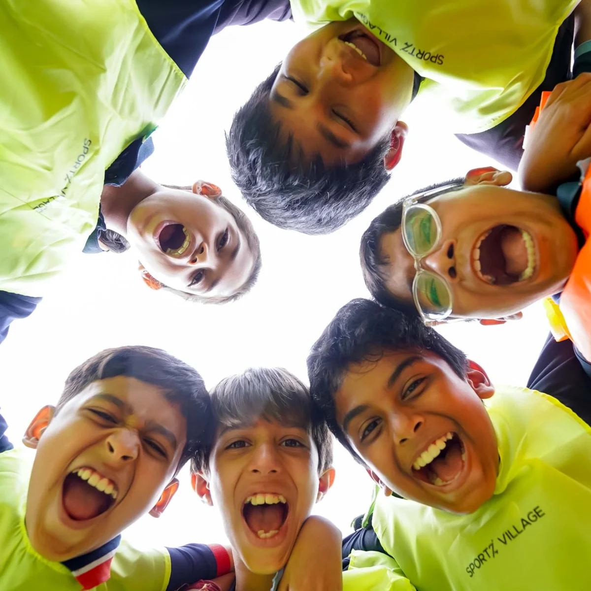 Students in neon yellow Sportz Village shirts huddle together laughing, viewed from below, showing team spirit and camaraderie