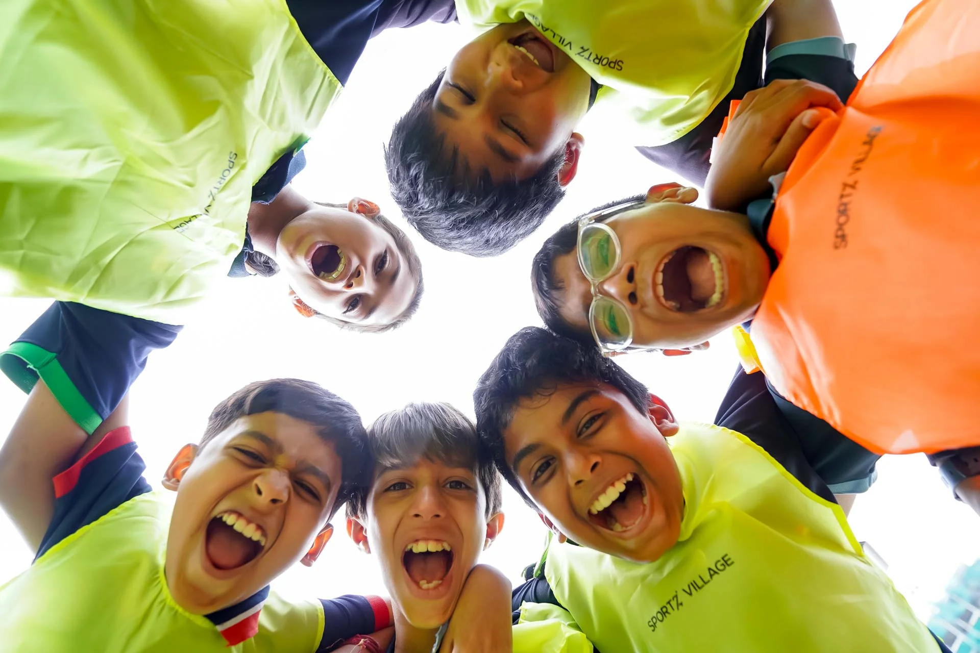 Students in neon yellow Sportz Village shirts huddle together laughing, viewed from below, showing team spirit and camaraderie